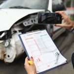 Woman photographs a broken car on smartphone and holds insurance documents in her hands. Damage assessment after car accident concept