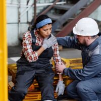 Mature foreman in workwear giving first aid to female subordinate in pain