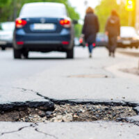 Damaged asphalt road with a large pothole in the daytime. The road surface shows cracks and unevenness. People walking and cars driving in the background. The image has a neutral tone and focuses on the damaged road surface. Suitable for articles on road maintenance, infrastructure, and construction.