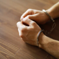 Hands in handcuffs rest on a wooden table, conveying a sense of confinement and seriousness. The metal cuffs are shiny against the wood grain.