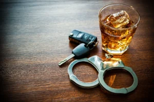 A glass of whiskey with ice, car keys, and handcuffs lie on a wooden table. The image conveys a cautionary message about drinking and driving.