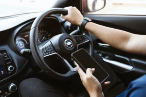 A person driving a car is using a smartphone with one hand on the wheel. The image highlights distracted driving, conveying a sense of danger.