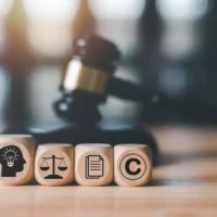 Wooden blocks depict icons of a brain, scales, document, and copyright symbol in focus. A gavel is blurred in the background, suggesting a legal theme.