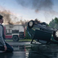 young-man-in-hoodie-kneels-on-the-road-looking-away-from-a-burning-overturned-car-after-a-traffic-accident